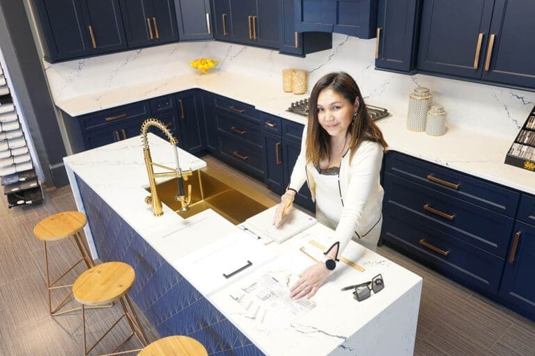 A smiling woman in a cream blazer stands at a white quartz island in a navy-cabinet kitchen, reviewing design samples.