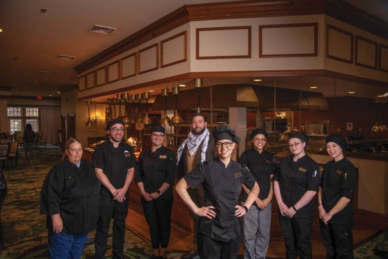 Eight restaurant staff in black chef's jackets stand smiling in a warmly lit dining room beneath a wooden ceiling soffit.