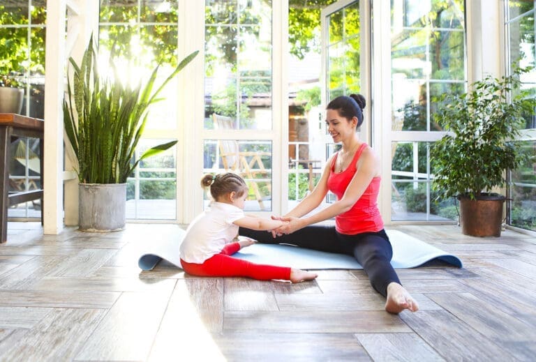 Young brunette woman with little daughter in a sporty uniform doing yoga on the terrace of a house by the garden