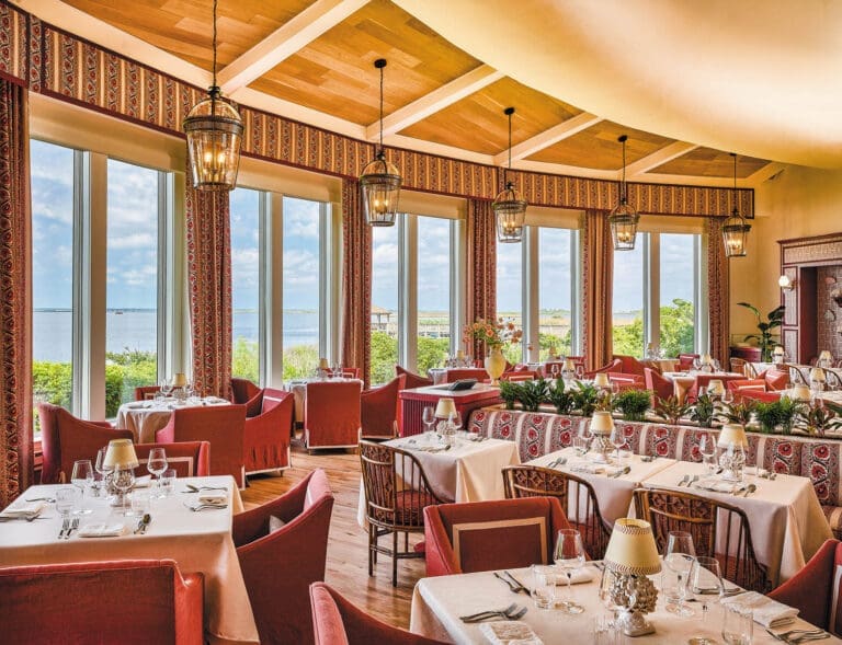 Elegant dining room with white-clothed tables, red upholstered chairs, lantern pendants, and large windows overlooking the water.