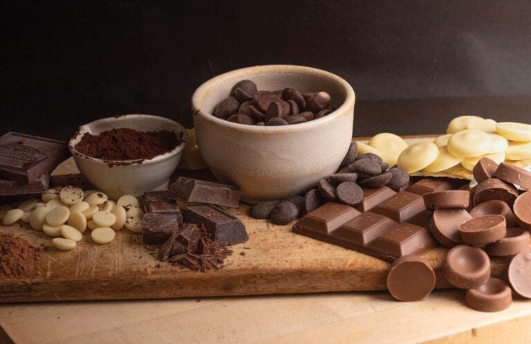 A rustic board displays dark chocolate chips in a bowl, cocoa powder, white and milk chocolate pieces, bars, and chunks.