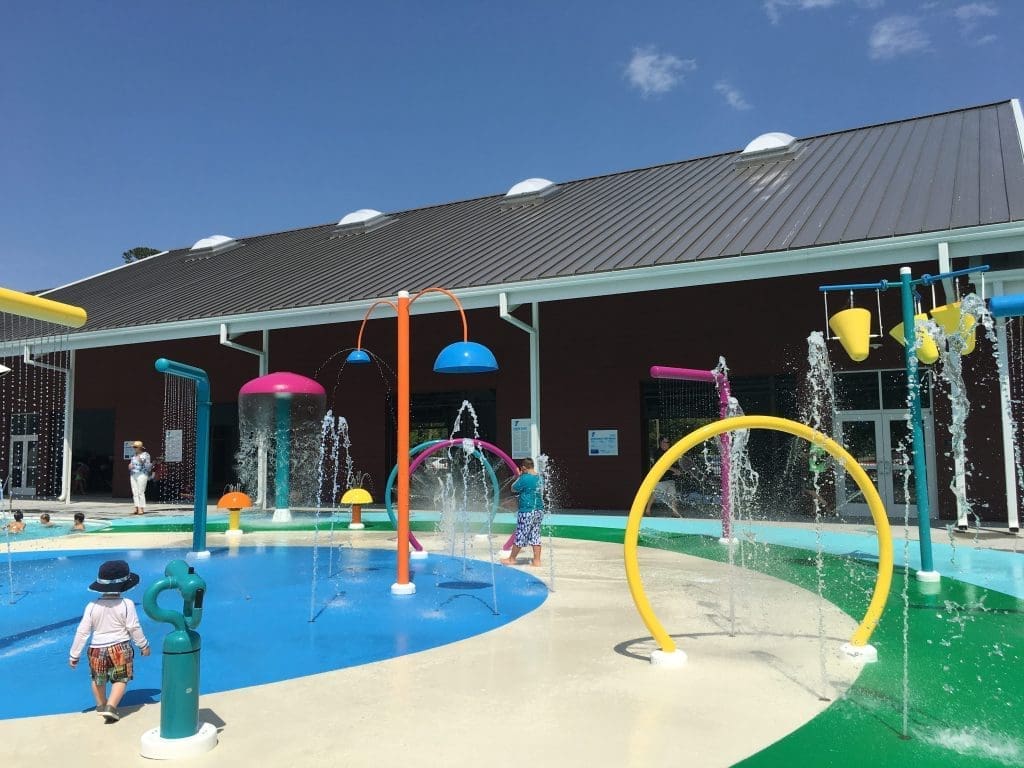 Colorful splash pad with water jets and arches, with children playing beside a dark red building under a blue sky.