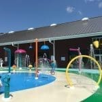 Colorful splash pad with water jets and arches, with children playing beside a dark red building under a blue sky.