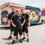 Three smiling men in black "Team Fat Kid" shirts and red caps stand before a wood-paneled food truck.
