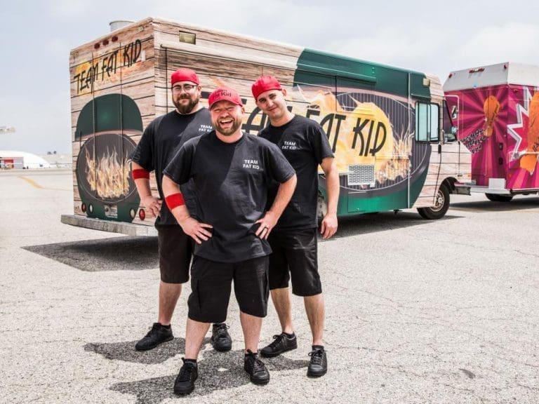 Three smiling men in black "Team Fat Kid" shirts and red caps stand before a wood-paneled food truck.