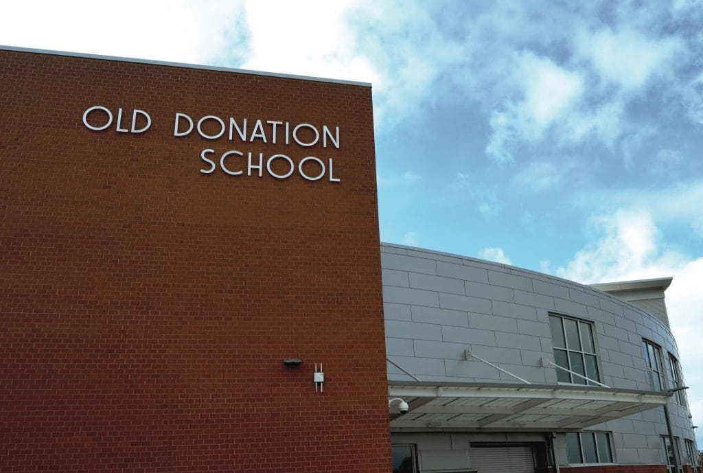 Brick and white-paneled exterior of Old Donation School with its name on the facade under a blue sky.