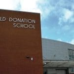 Brick and white-paneled exterior of Old Donation School with its name on the facade under a blue sky.