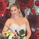 A smiling bride in a lace gown and veil holds a bouquet of white and pink blooms before a backdrop of large red paper flowers.