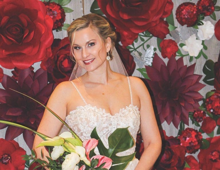 A smiling bride in a lace gown and veil holds a bouquet of white and pink blooms before a backdrop of large red paper flowers.