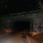 Nighttime view of a dark concrete underpass arch with graffiti-covered walls, with a wet road leading