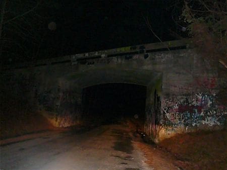 Nighttime view of a dark concrete underpass arch with graffiti-covered walls, with a wet road leading
