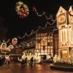 Illuminated clock tower and half-timbered buildings glow with holiday lights above a nighttime crowd in a Christmas village.