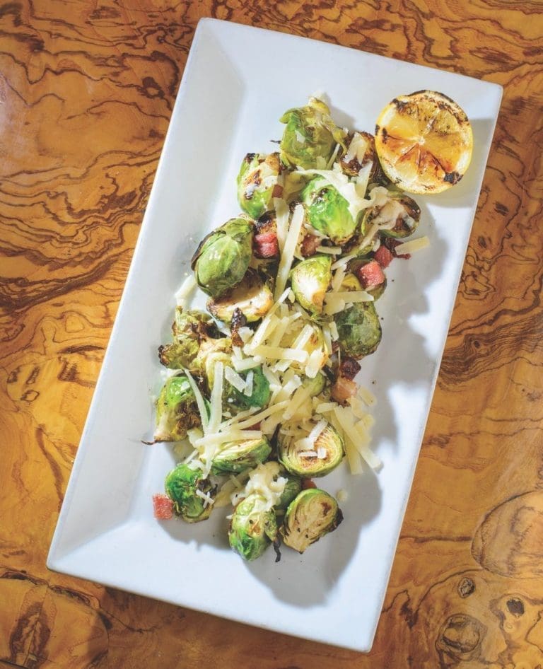 Charred Brussels sprouts with shaved Parmesan, diced pancetta, and a grilled lemon half on a white rectangular plate.