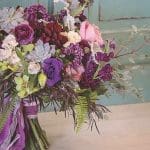 A lush bouquet of purple, pink and cream roses, succulents and greenery, tied with purple ribbon on a wooden table.