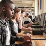 A young man in a plaid shirt works at a desktop computer beside classmates in a library-style computer lab.
