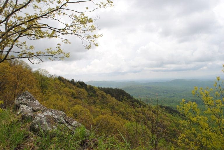 Spring overlook of rolling green mountain ridges under cloudy sky, with a large gray rock and budding trees in the foreground.