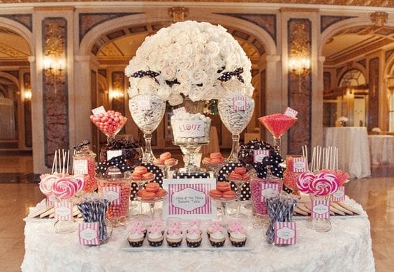 A candy buffet with white roses, pink and navy striped sweets, macarons, cupcakes, and lollipops on a white table.