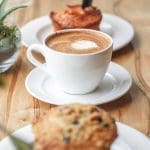 A white cup of latte with heart-shaped foam sits on a saucer, with blueberry muffins on plates nearby.