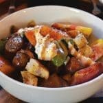 A cream-colored bowl of heirloom tomato salad with torn bread, green peppers, dollops of burrata, and drizzled dressing.