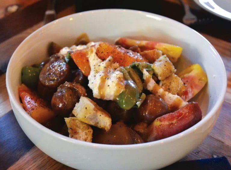 A cream-colored bowl of heirloom tomato salad with torn bread, green peppers, dollops of burrata, and drizzled dressing.