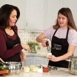 Two women in black "Cooking from the Heart" aprons prepare food in a white kitchen, one using a kitchen torch on a dessert.