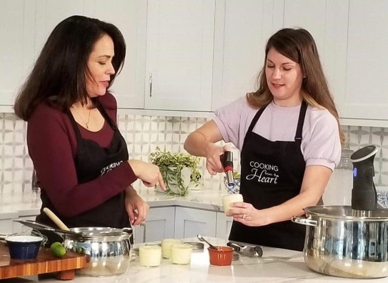 Two women in black "Cooking from the Heart" aprons prepare food in a white kitchen, one using a kitchen torch on a dessert.