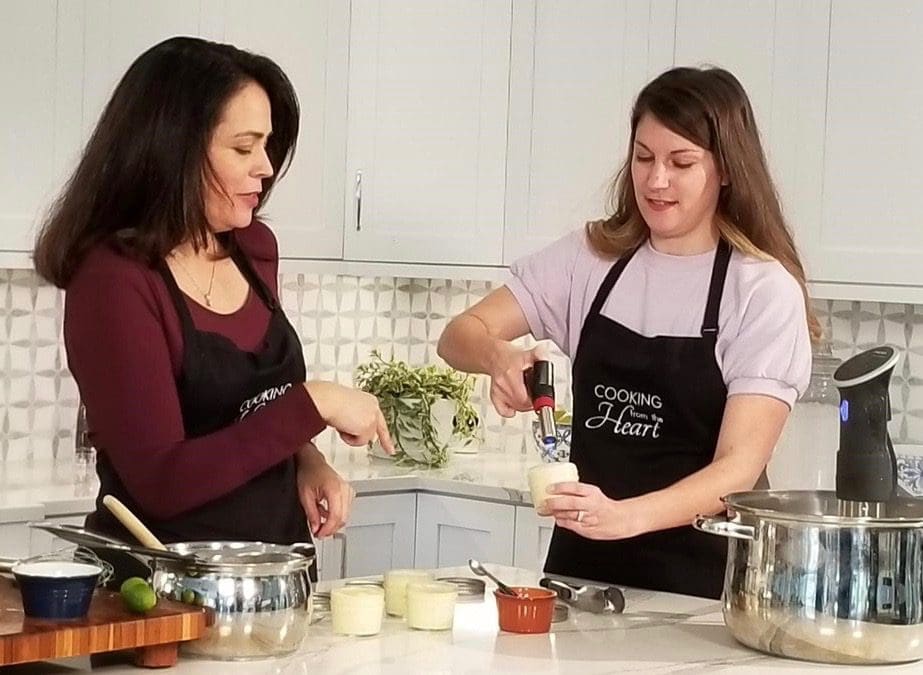 Two women in black "Cooking from the Heart" aprons prepare food in a white kitchen, one using a kitchen torch on a dessert.