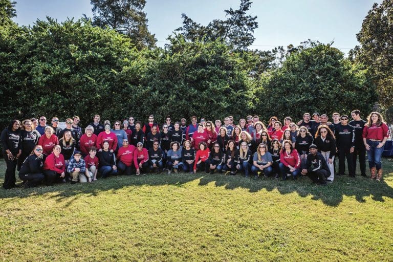 A large group of smiling women and a few men in red and black shirts posing outdoors on grass under trees.