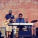 A five-piece band plays guitars, keyboard and drums in front of a red brick wall outdoors.