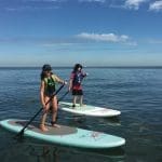 Two kids in life vests and caps paddleboard on calm, blue ocean water under a clear sky.