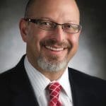 Smiling bald man with glasses, gray goatee, dark suit, striped shirt and red diamond-patterned tie against a dark backdrop.
