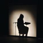 A silhouetted figure sits in a chair beside a microphone stand and music stand, lit against a glowing backdrop on a dark stage.