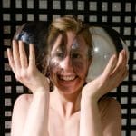 Smiling woman holds two clear glass spheres to her face, distorting her features against a black grid.