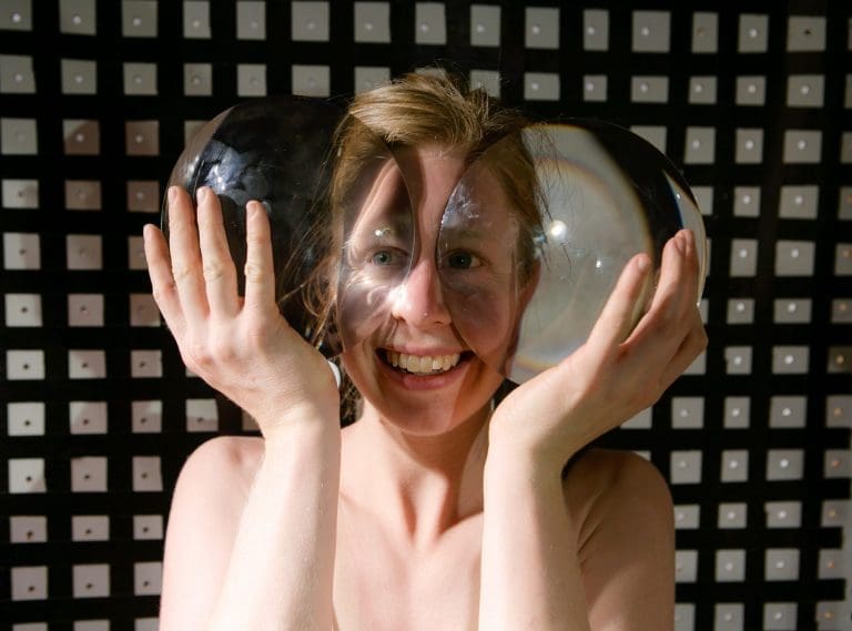 Smiling woman holds two clear glass spheres to her face, distorting her features against a black grid.