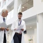 Two doctors in white coats walk through a bright hospital atrium, one reviewing paperwork while the other holds a clipboard.