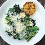 Overhead view of a white plate with charred broccolini and kale topped with shaved cheese, breadcrumbs and a grilled halved lemon.