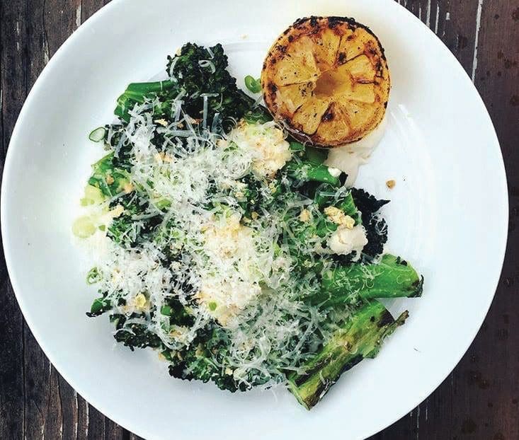 Overhead view of a white plate with charred broccolini and kale topped with shaved cheese, breadcrumbs and a grilled halved lemon.