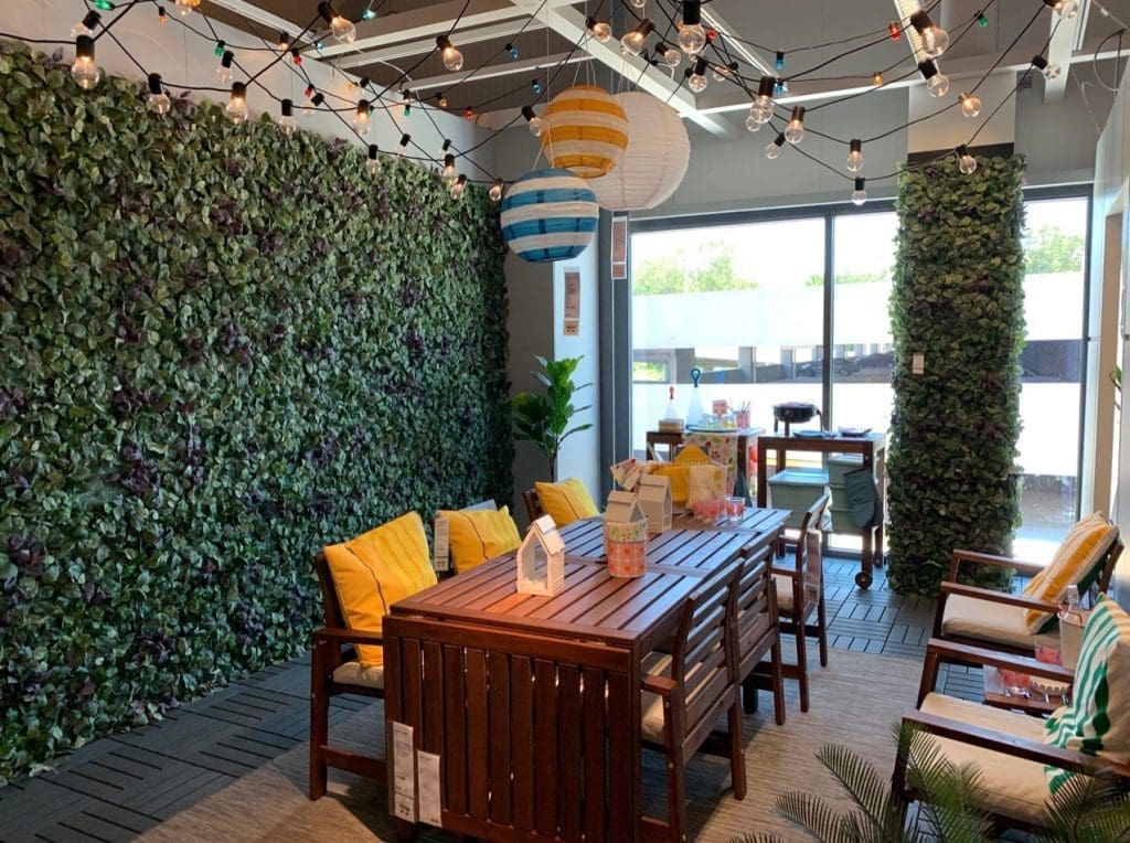 A furnished outdoor-style patio display with a greenery wall, string lights, wooden dining table, and yellow cushioned chairs.