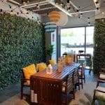 A furnished outdoor-style patio display with a greenery wall, string lights, wooden dining table, and yellow cushioned chairs.