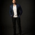 Bearded man in a navy blazer, white V-neck and black jeans, hands in pockets, against a dark backdrop.