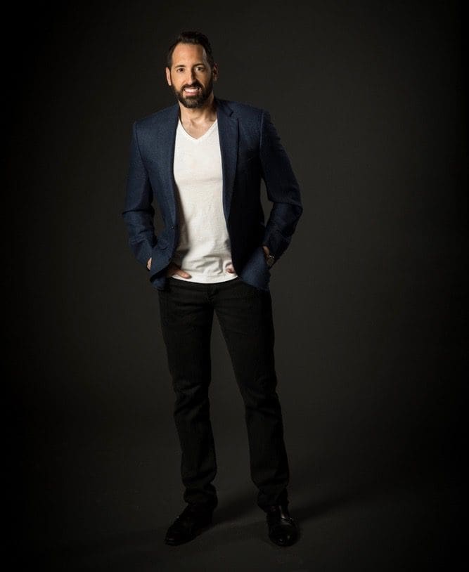Bearded man in a navy blazer, white V-neck and black jeans, hands in pockets, against a dark backdrop.