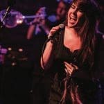 A woman with long dark hair sings passionately into a handheld microphone on a dimly lit stage, with a trumpet player behind her.