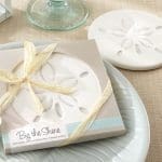 A white sand dollar stone coaster beside a gift box tied with yellow ribbon, labeled "By the Shore," on a pale plate.