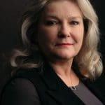 Studio portrait of Kate Mulgrew, a woman with wavy silver-blonde hair in a black jacket against a dark background.