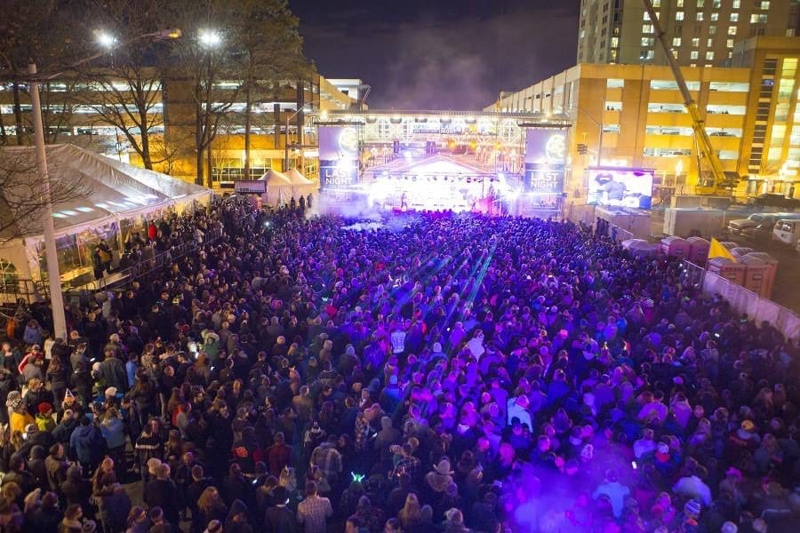 A large crowd fills a city street at night, facing a stage with purple lights, with signs reading "Last Night.