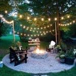 String lights hang over a brick patio with a stone fire pit, dark Adirondack chairs, and potted plants at dusk.