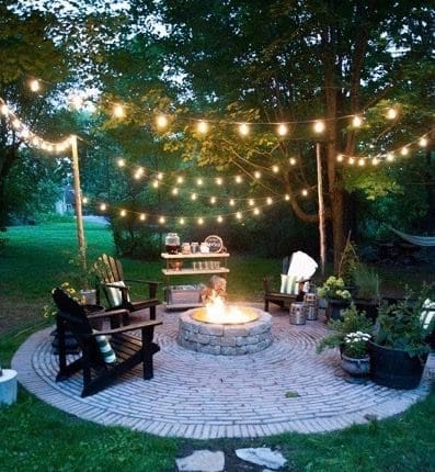 String lights hang over a brick patio with a stone fire pit, dark Adirondack chairs, and potted plants at dusk.