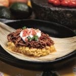 Shredded braised pork and sour cream on golden masa, served on a corn husk in a black dish, with diced tomato and cilantro.
