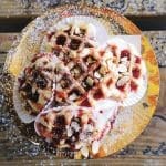 Three waffle-style pastries topped with raspberry jam, sliced almonds, and powdered sugar on a gold plate.