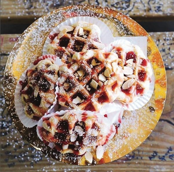 Three waffle-style pastries topped with raspberry jam, sliced almonds, and powdered sugar on a gold plate.